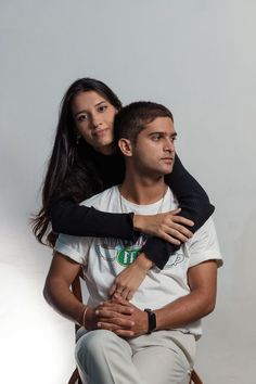 a man sitting next to a woman on top of a wooden chair in front of a white wall