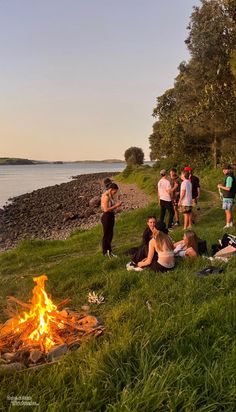 a group of people sitting around a fire pit on the grass near water and trees