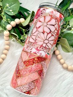 a pink and gold glitter tumbler next to green leaves on a white furnishing