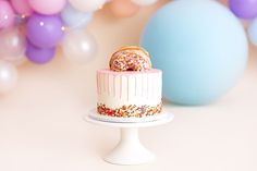 a white cake with sprinkles and a donut on top sits in front of balloons