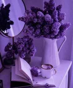 purple flowers are in a vase next to an open book and a cup of coffee