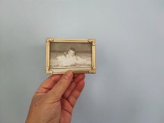 a hand holding an old fashioned photo in front of a gray wall with a gold frame