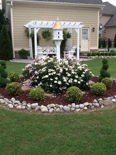 a white gazebo sitting in the middle of a flower bed next to a house