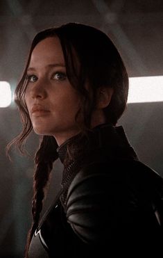 a woman with braids in a leather jacket looking off into the distance, standing in a dark room