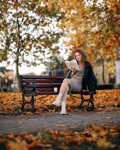 a woman sitting on a bench reading a book in an autumn park with leaves all over the ground