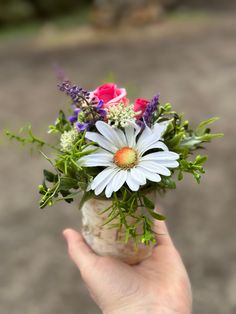 a hand holding a vase filled with flowers on top of a dirt road covered in grass