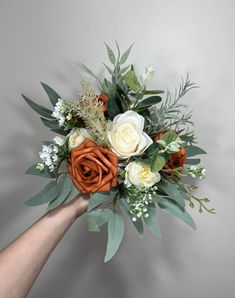 a person's hand holding a bouquet of white and orange flowers with greenery