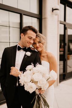 a bride and groom pose for a wedding photo in front of a building with large windows