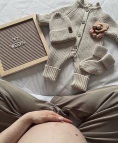 a pregnant woman laying on top of a bed next to a teddy bear and sweater