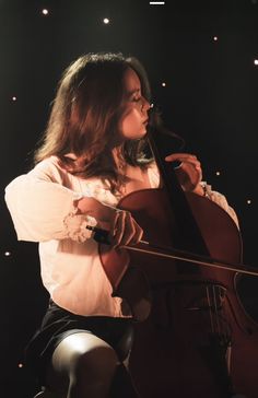 a woman in white shirt and black skirt playing cello on stage with lights behind her