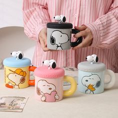 a person holding a coffee mug with snoopy faces on it and three other cups in front of them