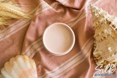 a cup of milk next to some sea shells on a pink and white striped blanket