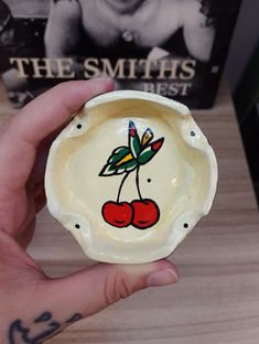 a hand holding a small bowl with cherries painted on the front and sides, in front of a book