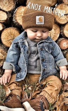 a baby sitting on top of a pile of logs wearing a knitted hat and denim jacket