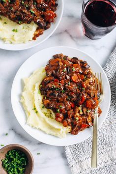 two white plates topped with mashed potatoes covered in meat and gravy next to a glass of wine