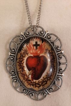 This silvertone necklace features a delightful mid-to-large-sized pendant with an openwork scalloped edge that gives it charm and character.  It is accented with a domed glass cameo with a strking image of the flaming red passionate Sacred Heart of Jesus/ The reverse has a textured finish and it measures 1 3/4 inches wide and 2 1/4 inches long. It includes a brooch fitting to allow it to be taken on and off the chain and worn as a pin if desired.  The chain is 24 inches and closes with a safety Sacred Heart Of Jesus, Heart Of Jesus, Sacred Heart, Scalloped Edge, Pendant Necklaces, Jewelry Necklace Pendant, Silver Tone, Jewelry Necklaces, Jesus