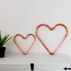 two heart shaped copper metal wall hooks on a white shelf next to a potted plant