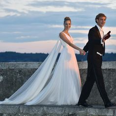 a bride and groom holding hands while walking along a stone wall in front of the ocean