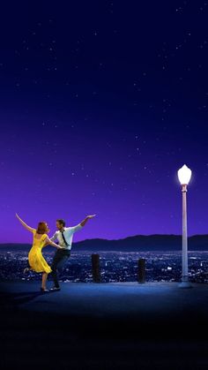 a man and woman dancing on top of a roof at night with the city lights in the background