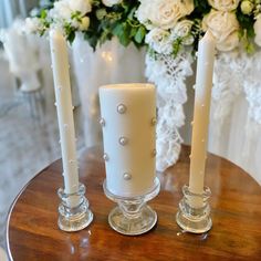 three white candles sitting on top of a wooden table next to flowers and vases