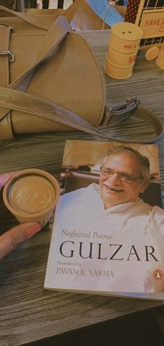 a person is holding a book near a purse and coffee cup on a table with other items