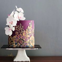 a purple and gold cake with white flowers on the top is sitting on a table