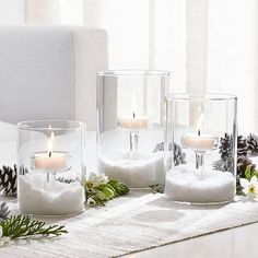 three glass vases filled with snow and lit candles on a table next to pine cones