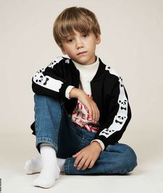 a young boy sitting on the ground wearing jeans and a black jacket with white letters