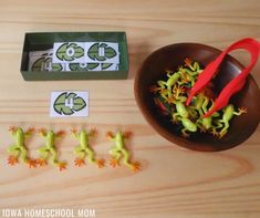 a wooden table topped with green plants and cut outs next to a bowl filled with red scissors