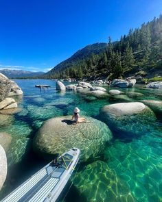 a person sitting on some rocks in the water next to a small boat and trees