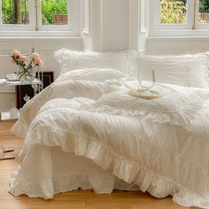 a bed with white ruffled bedspread and pillows in a room next to two windows