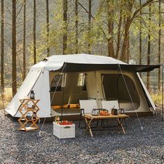 a tent is set up in the woods with chairs and tables on gravel ground next to it