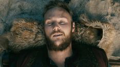 a man laying down with his eyes closed wearing a black shirt and furry coat over his shoulders