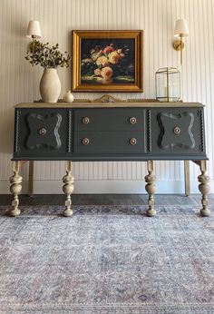 a painting on the wall above a black dresser with three drawers and two vases