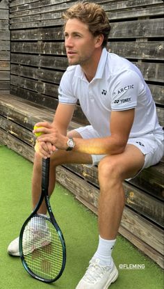 a man holding a tennis racquet on top of a grass covered court next to a wooden fence