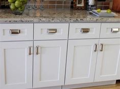 a kitchen with white cabinets and marble counter tops, green apples in a bowl on the island