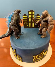 two godzilla figurines stand on top of a blue and yellow cake with city lights in the background