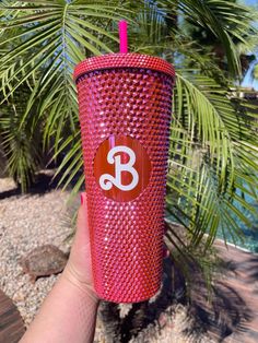 a person holding up a red cup with the letter b on it, in front of a palm tree