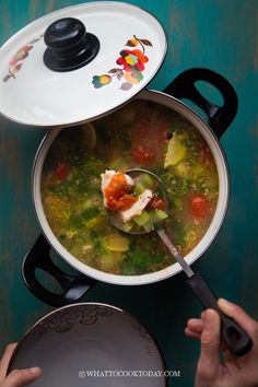 a person holding a spoon over a pot filled with soup on top of a table