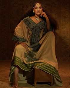 a woman sitting on top of a wooden bench wearing a dress and jewelry in front of a wall