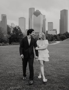 a man and woman walking in the grass with city buildings in the backgroud