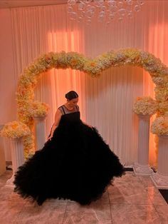 a woman in a black dress standing next to a white and yellow floral arch with flowers on it