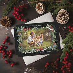 a christmas card with an image of two people sitting on a bench surrounded by holly and pine cones