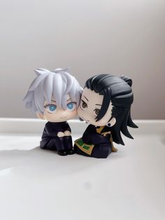 two anime figurines sitting next to each other on a white surface with blue eyes