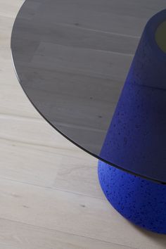 a round glass table with a blue base on a wooden floor in front of a white wall