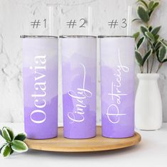 three purple tumblers with white writing on them sitting on a wooden tray next to a potted plant