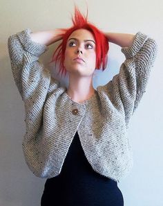 a woman with red hair wearing a gray cardigan and black dress standing in front of a white wall