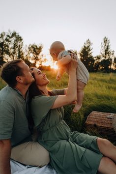 a man and woman are sitting on the grass with a baby in their lap, as the sun sets behind them