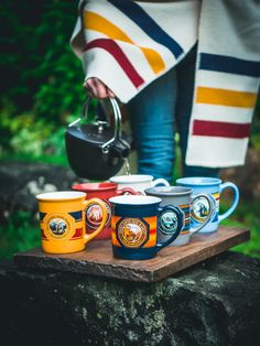 a person holding a tea kettle and cups on a wooden tray in front of some trees