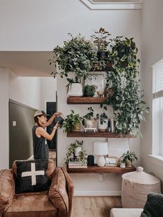 a woman standing in front of a living room with plants on the wall and shelves above her